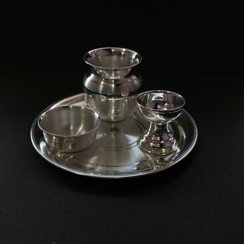 May include: A polished silver-toned metal set consisting of a circular tray, a small bowl, a rounded pot with a flared rim, and a pedestal-based cup. Each piece features a smooth, reflective surface and a simple, clean design for functional use.