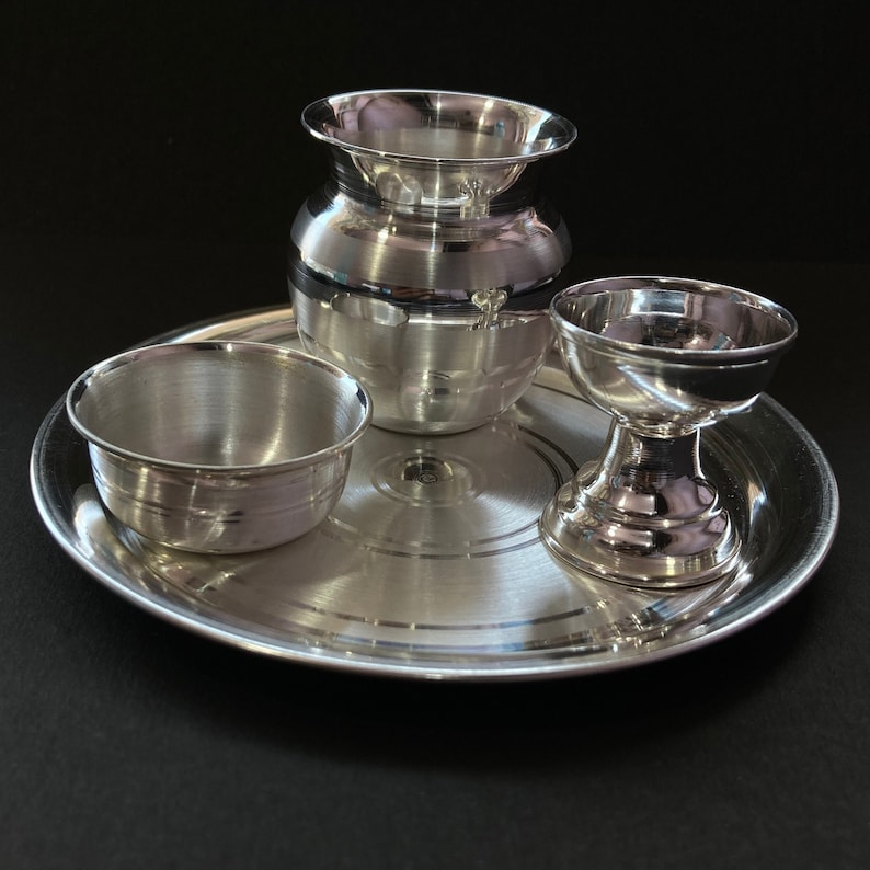 May include: A set of polished silver-coloured metal items consisting of a round tray, a small bowl, a rounded pot with a flared rim, and a pedestal-based cup. Each piece features a smooth, reflective surface with subtle concentric circular detailing.