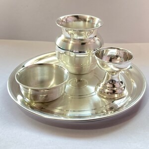 May include: A polished silver-coloured metal set consisting of a round tray, a small bowl, a tall pot with a flared rim, and a pedestal-based cup. Each piece features a smooth, reflective surface with subtle horizontal bands and a metallic finish.