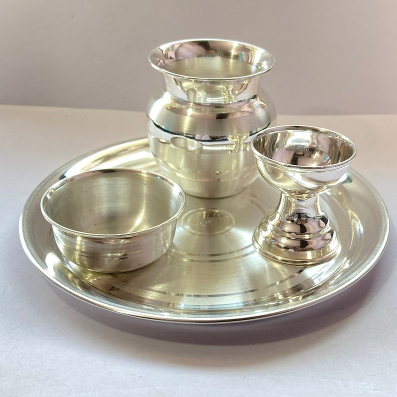 May include: A polished silver-coloured metal set consisting of a round tray, a small bowl, a tall pot with a flared rim, and a pedestal-based cup. Each piece features a smooth, reflective surface with subtle horizontal bands and a metallic finish.