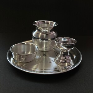 May include: A polished silver-toned metal set consisting of a round tray, a small bowl, a wide-mouthed pot, and a pedestal cup. Each piece features a smooth, reflective surface with subtle concentric circular detailing on the base of the tray.