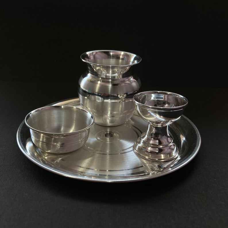 May include: A polished silver-toned metal set consisting of a round tray, a small bowl, a wide-mouthed pot, and a pedestal cup. Each piece features a smooth, reflective surface with subtle concentric circular detailing on the base of the tray.