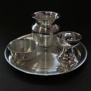 May include: A polished silver-toned metal set consisting of a round tray, a small bowl, a rounded pot with a wide rim, and a pedestal-based cup. Each piece features a smooth, reflective surface with subtle concentric circular detailing on the tray.