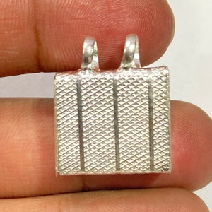 May include: Silver pendant with a square shape and a textured surface. The pendant has two loops at the top for attaching a chain.