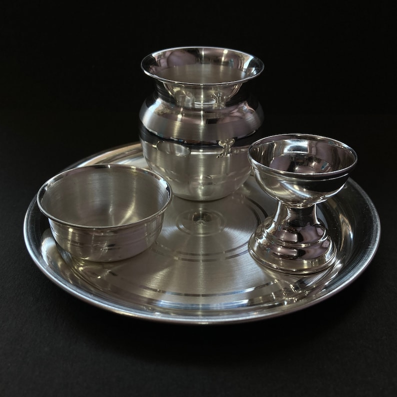 May include: A polished silver-toned metal set consisting of a round tray, a small bowl, a wide-mouthed pot, and a pedestal cup. Each piece features a smooth, reflective surface with subtle concentric circular detailing on the tray base.