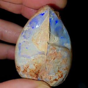 May include: A teardrop-shaped opal gemstone with a rough, textured exterior. The stone features a mix of colors, including blue, white, and brown, with flashes of iridescent color. The opal is held between fingers against a dark background.