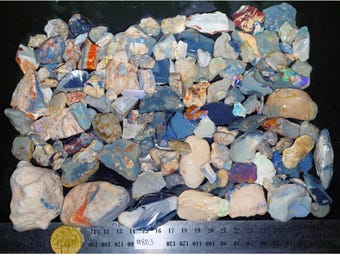 Op de afbeelding: Een verzameling ruwe opaalstenen in verschillende vormen en maten, met een reeks kleuren, waaronder blauw, wit en oranje. Een liniaal met centimeter maten staat onderaan, samen met een munt. De tekst "#803" is zichtbaar.