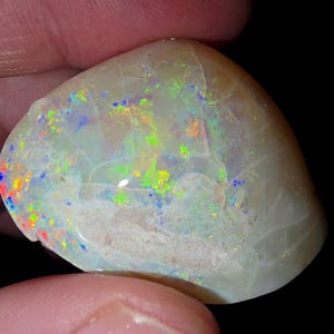 May include: A polished opal gemstone with a creamy white base color. The opal displays vibrant flashes of color, including blue, green, yellow, and red, creating a play-of-color effect. The stone has a smooth, rounded shape.