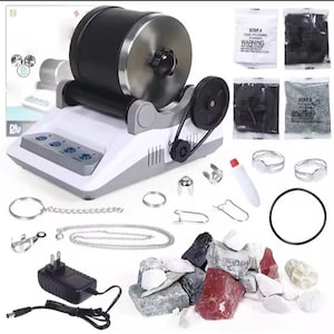 Op de afbeelding: Een rots-tumbler kit met een zwarte en zilveren trommel, een witte basis met bedieningsknoppen en diverse accessoires. Inclusief zakjes polijstkorrel, sieradenonderdelen, een voedingsadapter en een selectie ruwe edelstenen.