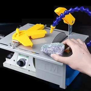 Pode incluir: Uma pequena serra lapidária cinza com uma lâmina circular, proteções amarelas e um tubo de água azul flexível. Uma mão segura uma rocha multicolorida perto da lâmina. A serra tem um botão de controle e uma base cinza e azul.