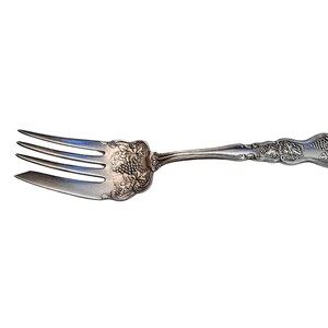 May include: Silver fork with three tines and a decorative handle. The handle features an ornate design with grape clusters and leaves. The fork is likely vintage or antique, and is set against a white background.