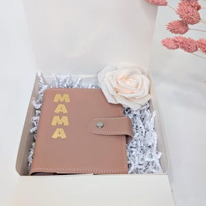 May include: A pink leather journal with the word "MAMA" in gold lettering. The journal is in a white gift box with white shredded paper and a single pink rose.