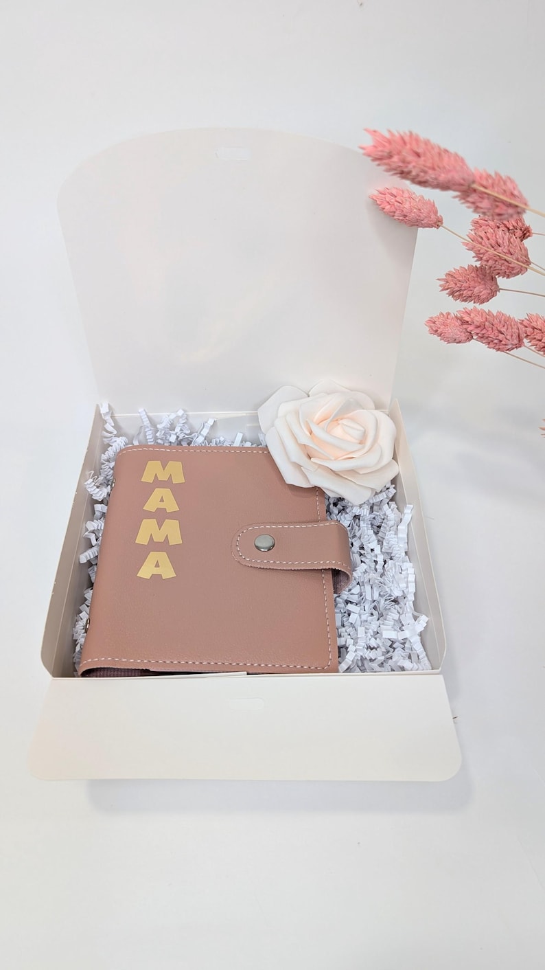 May include: A pink leather journal with the word "MAMA" in gold lettering. The journal is in a white gift box with white shredded paper and a single pink rose.
