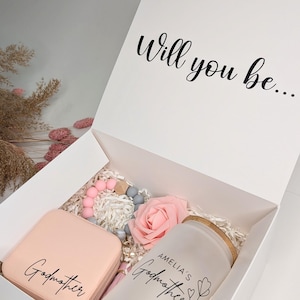 Pu&ograve; includere: Una scatola regalo bianca contenente un braccialetto in silicone rosa e grigio, una rosa rosa, una scatola per gioielli rosa con la scritta "Madrina" e un barattolo di vetro smerigliato con la scritta "Madrina di Amelia".