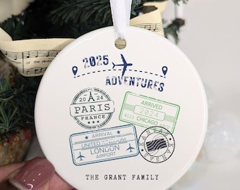 May include: A white ceramic Christmas ornament with travel-themed stamps and the text "2025 Adventures." Stamps include "Paris, France," "London Airport," and "Chicago." The ornament is held by a hand and has a white ribbon for hanging.