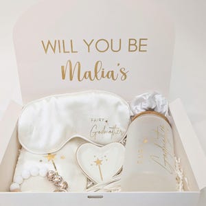 Puede incluir: Caja de regalo blanca con letras doradas que dicen "Will you be Malia's Fairy Godmother?" La caja contiene una máscara para dormir de satén blanco, un plato blanco en forma de corazón con un diseño de estrella dorada y varita, un vaso blanco con letras doradas que dicen "Fairy Godmother" y un collar de cuentas blancas con detalles dorados.