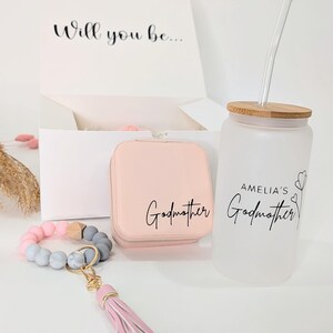Pu&ograve; includere: Un set regalo per una madrina. Il set include una scatola per gioielli rosa con la scritta "Madrina" su di essa, un braccialetto di perline rosa e grigio con un portachiavi a nappa rosa e una bottiglia d'acqua in vetro smerigliato con tappo in bamb&ugrave; e cannuccia. La bottiglia d'acqua ha il testo "Madrina di Amelia" su di essa.