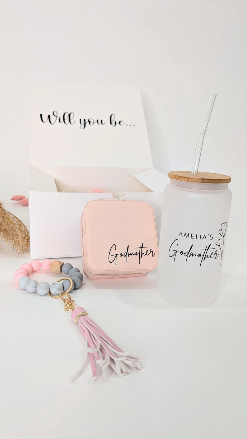 Pu&ograve; includere: Un set regalo per una madrina. Il set include una scatola per gioielli rosa con la scritta "Madrina" su di essa, un braccialetto di perline rosa e grigio con un portachiavi a nappa rosa e una bottiglia d'acqua in vetro smerigliato con tappo in bamb&ugrave; e cannuccia. La bottiglia d'acqua ha il testo "Madrina di Amelia" su di essa.