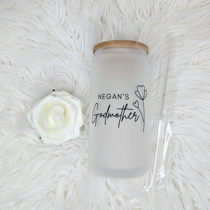 Pu&ograve; includere: Un barattolo di vetro bianco smerigliato con un coperchio in bamb&ugrave; e una scritta nera che recita "Negan's Godmother" con un semplice disegno di un fiore. Il barattolo &egrave; su una superficie bianca e soffice.