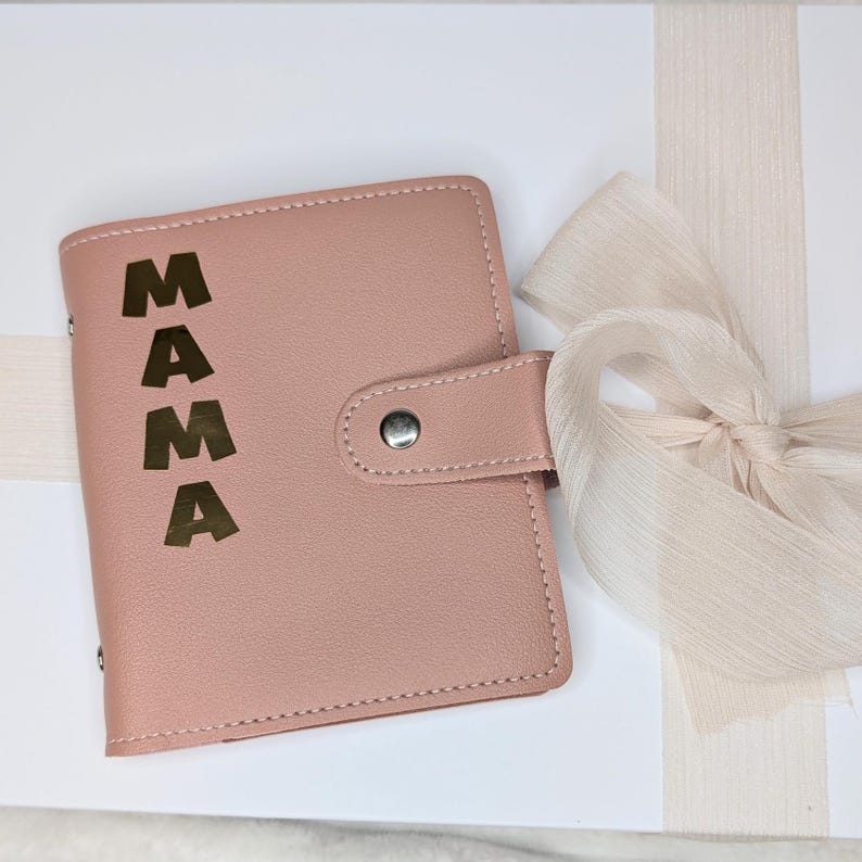 May include: A blush pink leather-bound journal with a gold foil inscription that reads "MAMA". The journal is wrapped in a white box with a cream-colored ribbon.