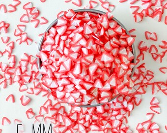 May include: A bowl filled with red and white strawberry-shaped sprinkles, with additional sprinkles scattered around the bowl on a white surface. The sprinkles are 5 mm in size. The text "Original Six Designs and Crafts" is visible below the bowl.