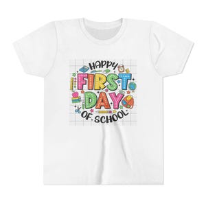 Puede incluir: Una camiseta blanca de manga corta con un gráfico central sobre un fondo de cuadrícula. El diseño incluye el texto "HAPPY FIRST DAY OF SCHOOL" en letras coloridas y punteadas, rodeado de ilustraciones de un globo terráqueo, mochila, libros y útiles.