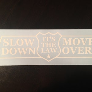 Puede incluir: Calcomanía de vinilo blanco con el texto "Slow Down It's The Law. Move Over" en forma de escudo. La calcomanía está sobre un fondo azul claro.