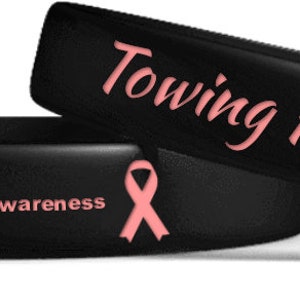Puede incluir: Dos pulseras de silicona negras con diseños de cintas rosas. Una pulsera dice "Towing For A Cure". La otra pulsera dice "Breast Cancer Awareness."