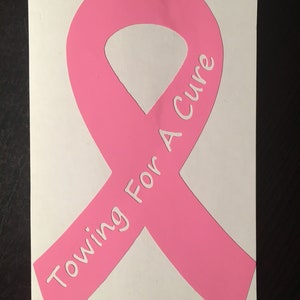 Puede incluir: Cinta rosa con el texto "Towing For A Cure" en letras blancas. La cinta es un símbolo de concienciación sobre el cáncer de mama.