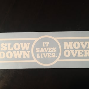 Puede incluir: Calcomanía de vinilo blanco con el texto "Slow Down It Saves Lives. Move Over" sobre un fondo azul claro.