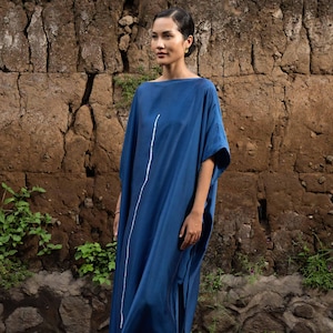 May include: A long, flowing, dark blue dress with a boat neckline and short sleeves. A vertical white line runs down the front of the dress. The model is standing in front of a textured brown stone wall, with purple flower petals on the ground.