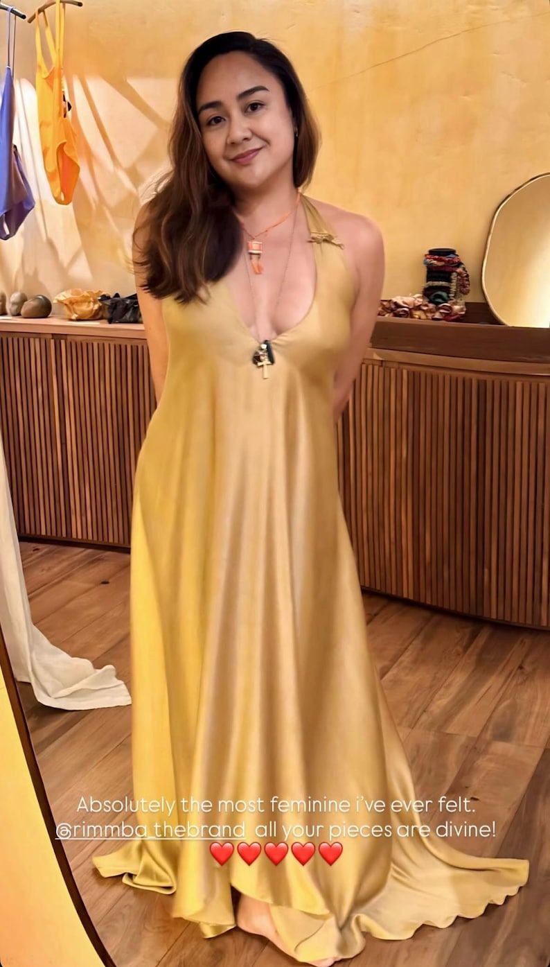 Op de afbeelding: Een lange, vloeiende, goudgele jurk met een halternek. De jurk heeft een lage halslijn en een zijdeachtige uitstraling. De afbeelding bevat tekst die luidt: "Absolutely the most feminine I've ever felt."