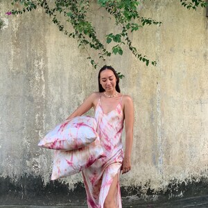 Puede incluir: Una mujer lleva un vestido maxi de color rosa y blanco con estampado tie-dye y una abertura lateral. Est&aacute; sosteniendo dos almohadas a juego de color rosa y blanco con estampado tie-dye.