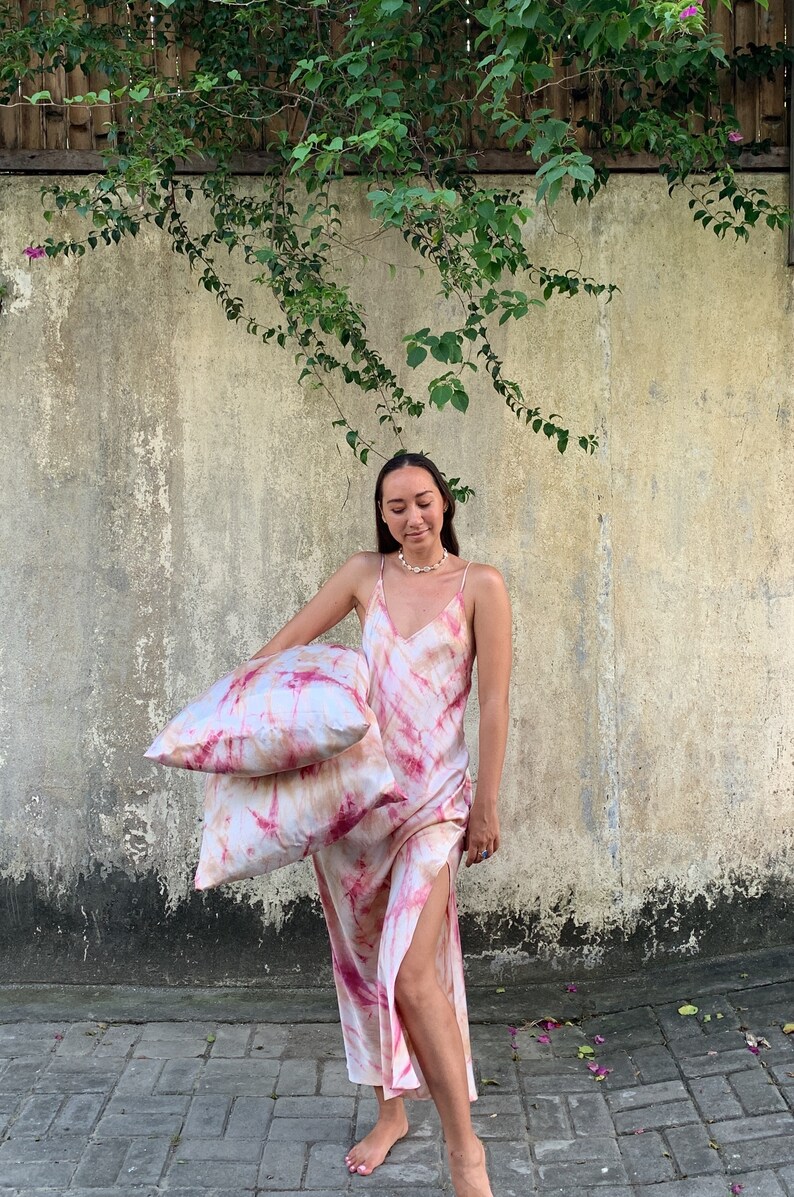 Puede incluir: Una mujer lleva un vestido maxi de color rosa y blanco con estampado tie-dye y una abertura lateral. Est&aacute; sosteniendo dos almohadas a juego de color rosa y blanco con estampado tie-dye.