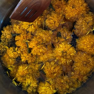 Op de afbeelding: Een bovenaanzicht van een roestvrijstalen pot gevuld met heldergele goudsbloembloemen. Een houten spatel rust op de rand van de pot. De bloemen zijn dicht opeengepakt, waardoor een levendig, gestructureerd oppervlak ontstaat.