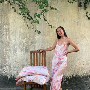 Puede incluir: Dos fundas de almohada de color rosa y blanco con estampado tie-dye apiladas en una silla de madera. Una persona con un vestido de color rosa y blanco con estampado tie-dye se encuentra detr&aacute;s de la silla.