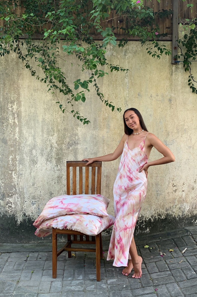 Puede incluir: Dos fundas de almohada de color rosa y blanco con estampado tie-dye apiladas en una silla de madera. Una persona con un vestido de color rosa y blanco con estampado tie-dye se encuentra detr&aacute;s de la silla.