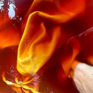 Op de afbeelding: Close-up van stof ondergedompeld in een rood-oranje vloeistof. De stof is helder geel-oranje, met plooien en vouwen die textuur cre&euml;ren. De vloeistof lijkt kleurstof te zijn, wat wijst op een textielverfproces.