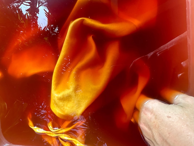 Op de afbeelding: Close-up van stof ondergedompeld in een rood-oranje vloeistof. De stof is helder geel-oranje, met plooien en vouwen die textuur cre&euml;ren. De vloeistof lijkt kleurstof te zijn, wat wijst op een textielverfproces.