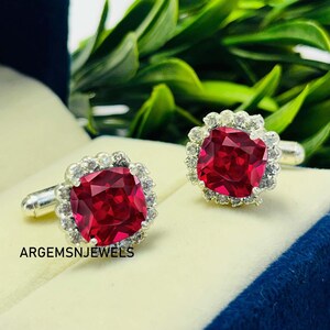 Peut inclure: Une paire de boutons de manchette en argent avec de grandes pierres rouges carrées, entourées d'un halo de petites pierres claires. Les boutons de manchette sont présentés sur une surface claire, avec un fond de feuillage vert. Le texte "ARGEMSNJEWELS" est visible.