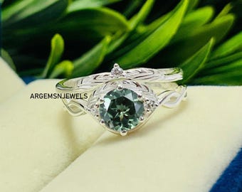 May include: A silver engagement ring and wedding band set. The engagement ring features a round, green gemstone surrounded by small, clear stones. The band has a delicate, swirling design. The text "ARGEMSNJEWELS" is visible.