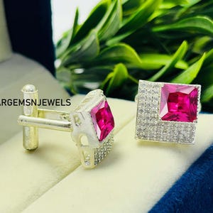 Peut inclure: Boutons de manchette argentés avec une pierre précieuse rose vif de forme carrée, entourée de petites pierres claires. Les boutons de manchette ont une forme rectangulaire. Le texte "ARGEMSNJEWELS" est visible.