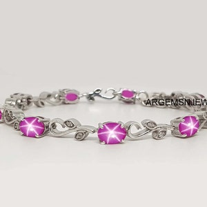 Puede incluir: Una pulsera de plata con zafiros estrella rosa y pequeños diamantes engastados en un diseño de hojas. La pulsera tiene un cierre de broche. El texto "ARGEMSNJEWELS" es visible en la imagen.