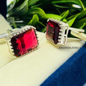 May include: A pair of silver cufflinks featuring large, rectangular, deep red gemstones. The gemstones are set in a silver frame with a decorative edge. The cufflinks are displayed on a light blue surface.