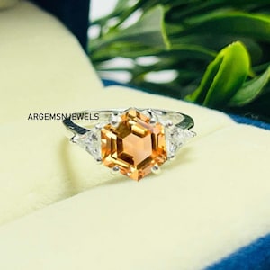 Può includere: Un anello d'argento con una grande pietra preziosa esagonale arancione circondata da tre piccole pietre preziose triangolari trasparenti. L'anello è in una scatola bianca con bordo blu.