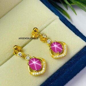 May include: A pair of gold earrings with a star ruby gemstone. The earrings have a gold setting with small, clear gemstones around the ruby. The ruby is a deep pink color with a star-like pattern. The earrings are displayed in a blue jewelry box.