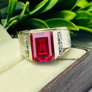 May include: A gold ring with a large, emerald-cut ruby gemstone and smaller, clear gemstones set in the band. The ring is in a small, brown box.