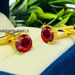 Peut inclure: Une paire de boutons de manchette dorés avec de grosses pierres précieuses rondes et rouge foncé. Les boutons de manchette sont présentés sur une surface bleu clair et blanche, avec un feuillage vert en arrière-plan. Le texte "ARGEMSN JEWELS" est visible.