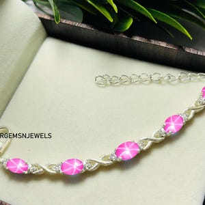 Könnte beinhalten: Ein silbernes Armband mit einem gedrehten Design und fünf rosa Stern-Saphiren. Das Armband hat einen Karabinerverschluss und ein kleines Schild mit dem Text "ARGEMSNJEWELS".