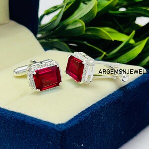 Peut inclure: Paire de boutons de manchette argentés avec des pierres rouges profondes de forme rectangulaire. Chaque bouton est bordé de petites pierres claires. Les boutons de manchette sont présentés dans un écrin en velours bleu. Le texte "ARGEMSNJEWELS" est visible.